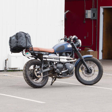 Blue Triumph Scrambler motorcycle with custom exhaust resting on it's kick stand with a waterproof duffle bag on the rear rack.